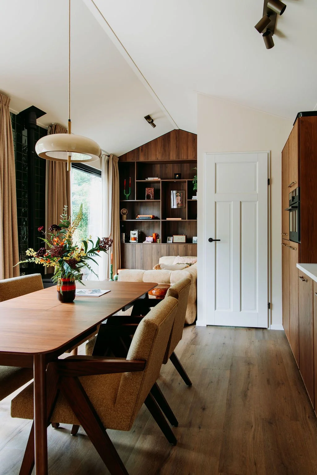Interieur van een modern eethoek met een houten tafel, beige stoelen en een vaas met bloemen. Achterin een zithoek met een ingebouwde houten boekenkast en een beige bank.