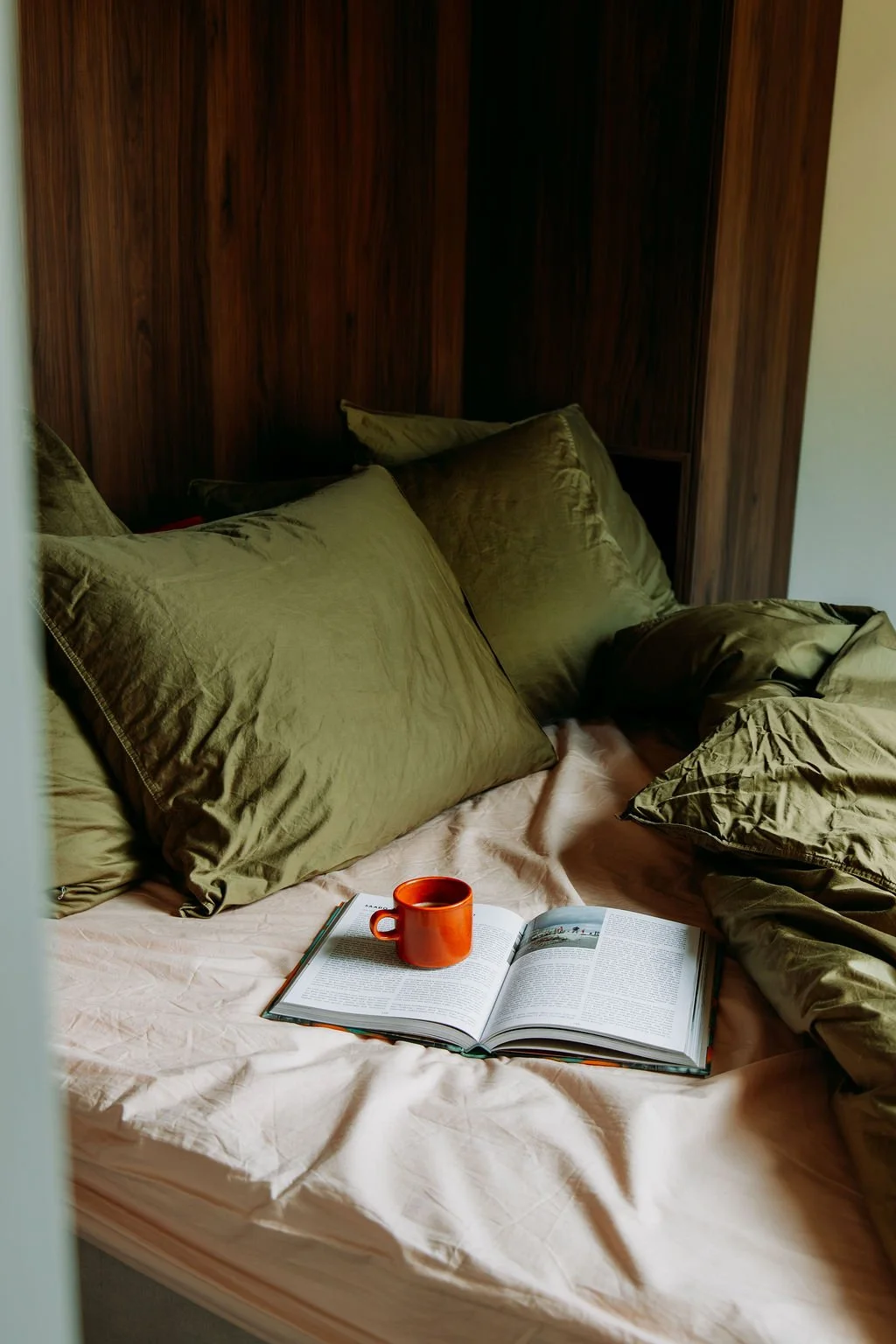 Een bed met grote groene kussens, een open boek en een oranje mok op een roze beddengoed.