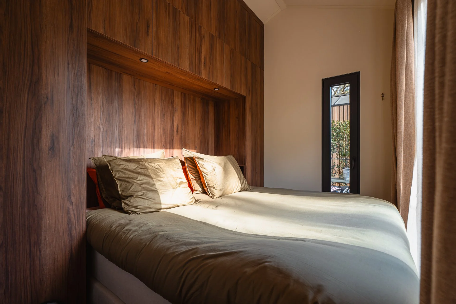 Een slaapkamer met een dubbelbed, beddengoed en kussens, een houten hoofdeinde en wand, grote raam met gordijnen, en een deur naar een balkon buiten.