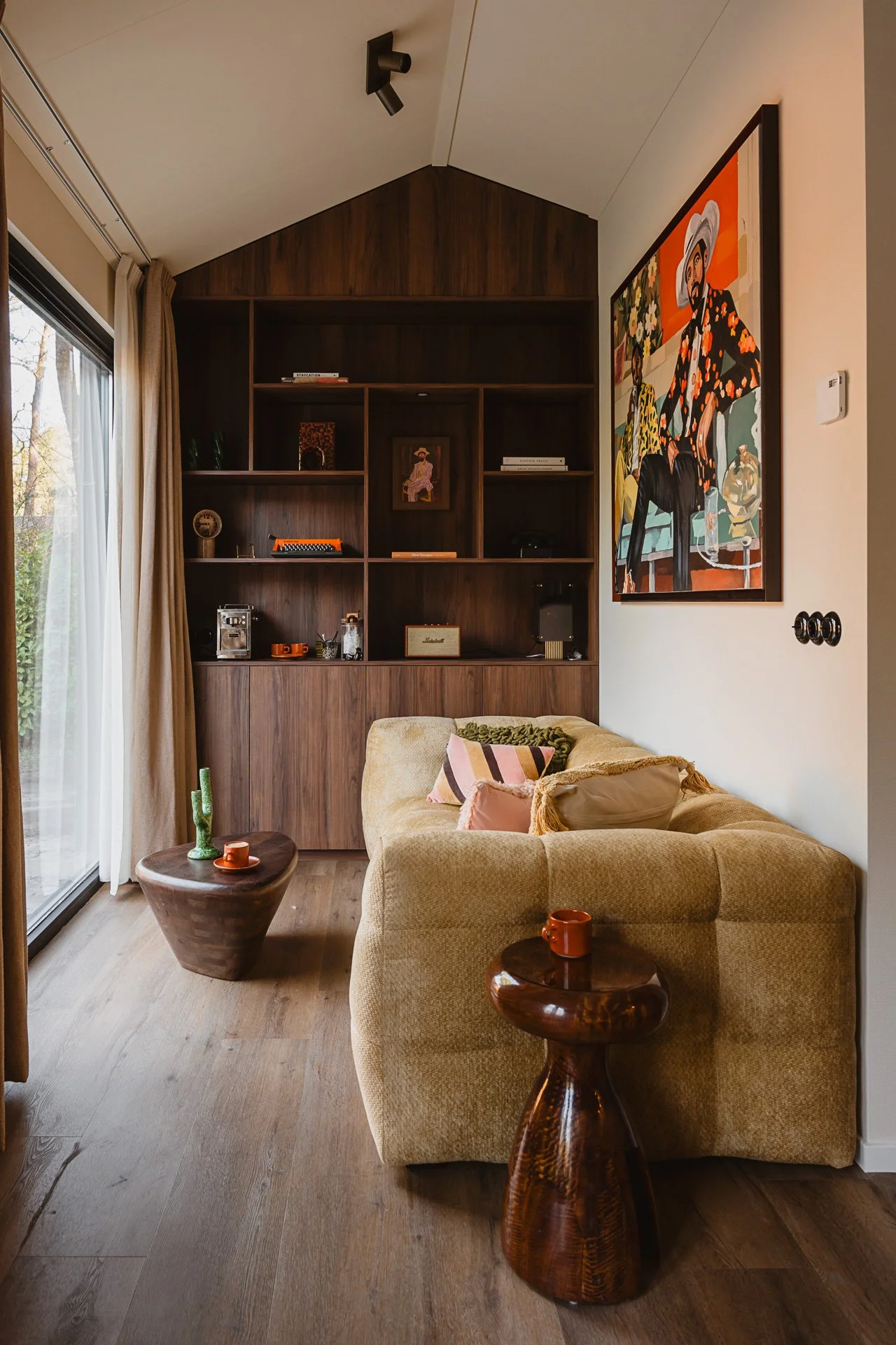 Gezellige woonkamer met beige bank, houten tafeltjes, grote kunst aan de muur, houten plankenwand en grote raam met gordijnen, herfstbladeren zichtbaar buiten.