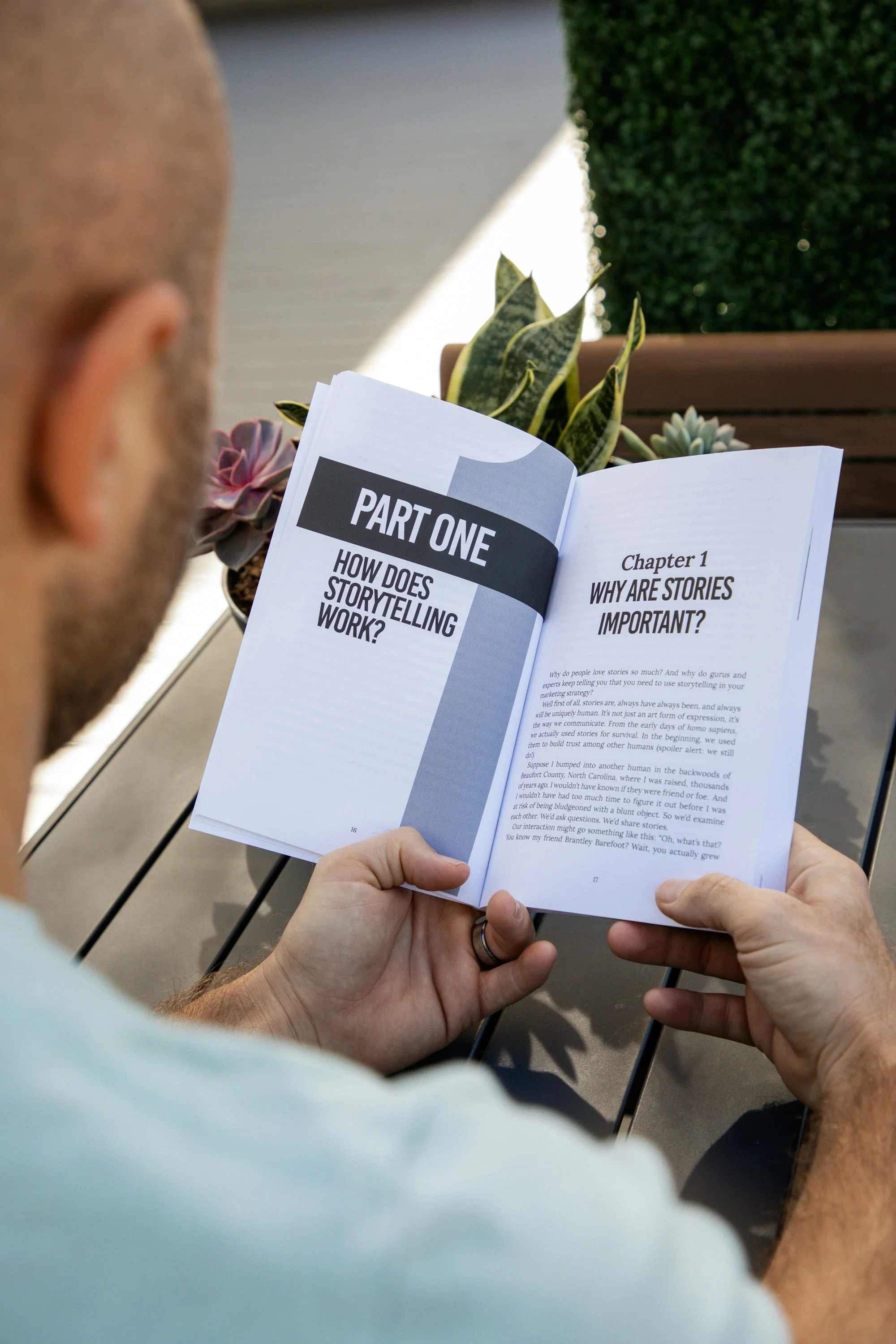 A person reading a booklet titled 'Part One: How Does Storytelling Work?' outdoors, with a table, potted succulents, and a green hedge in the background.