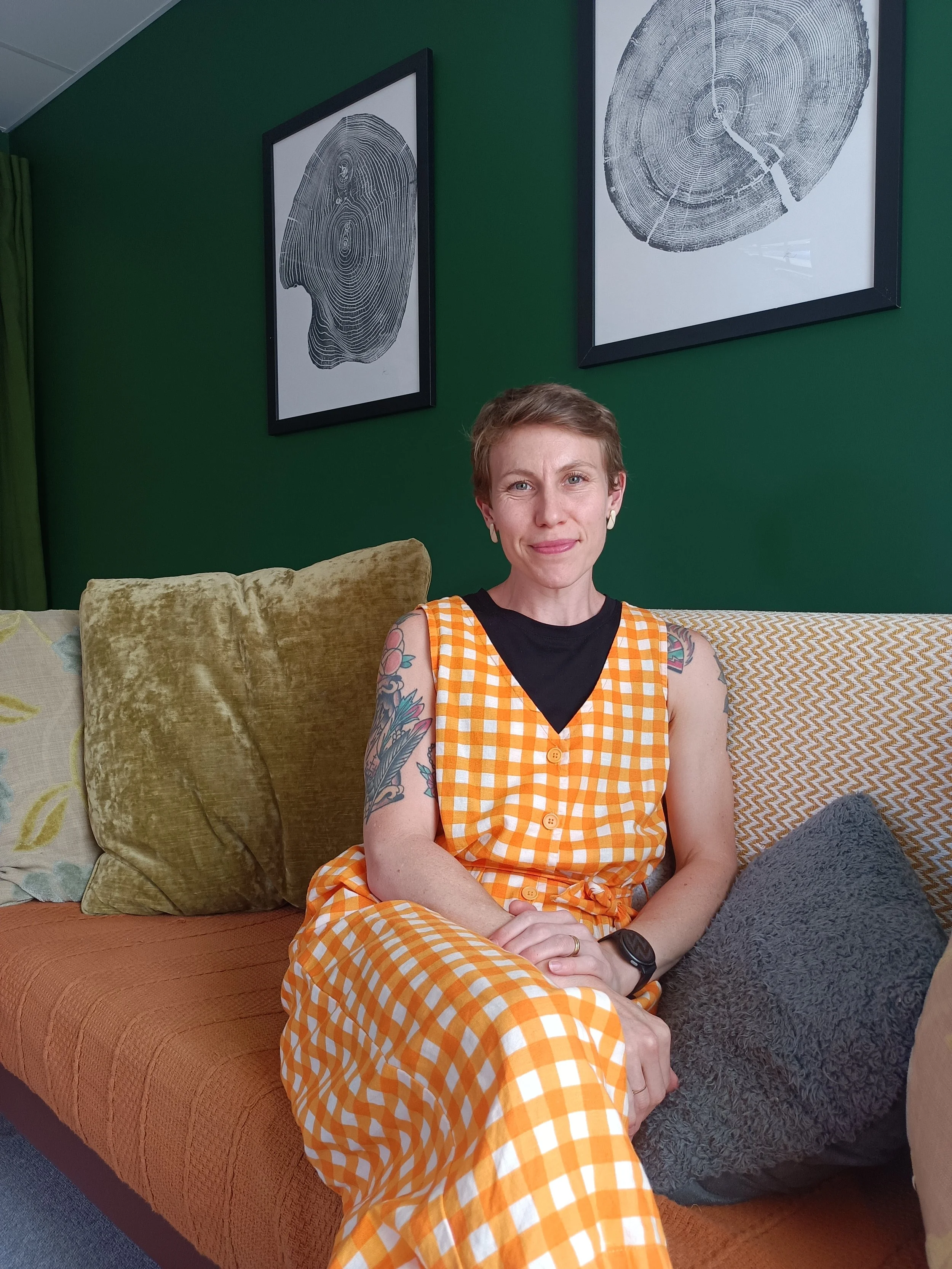 A woman with short hair, tattoos on arms, wearing an orange and white checkered dress over a black T-shirt, sitting on a tan couch with various pillows, in a room with green walls and framed black-and-white artwork.