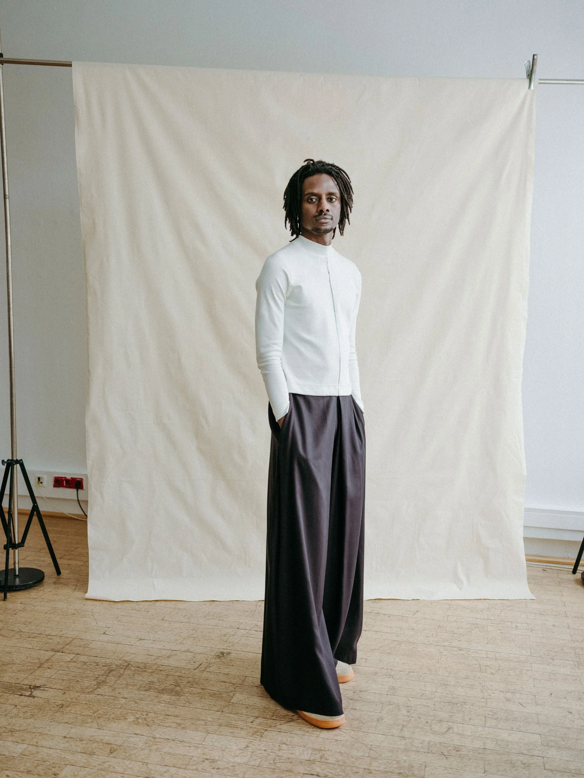 Eine Person mit Dreadlocks steht vor einem sichtbaren Studioaufbau, der den Hintergrund bildet. Sie trägt ein helles Oberteil und eine sehr weit geschnittene, dunkle Hose von studio.HIER.