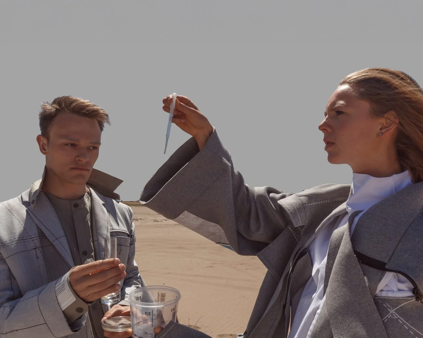 Zwei Menschen in Anzügen in einer Wüstenlandschaft, einer hält einen Teststab und der andere trägt eine Pipette. Sie scheinen eine wissenschaftliche Untersuchung durchzuführen.