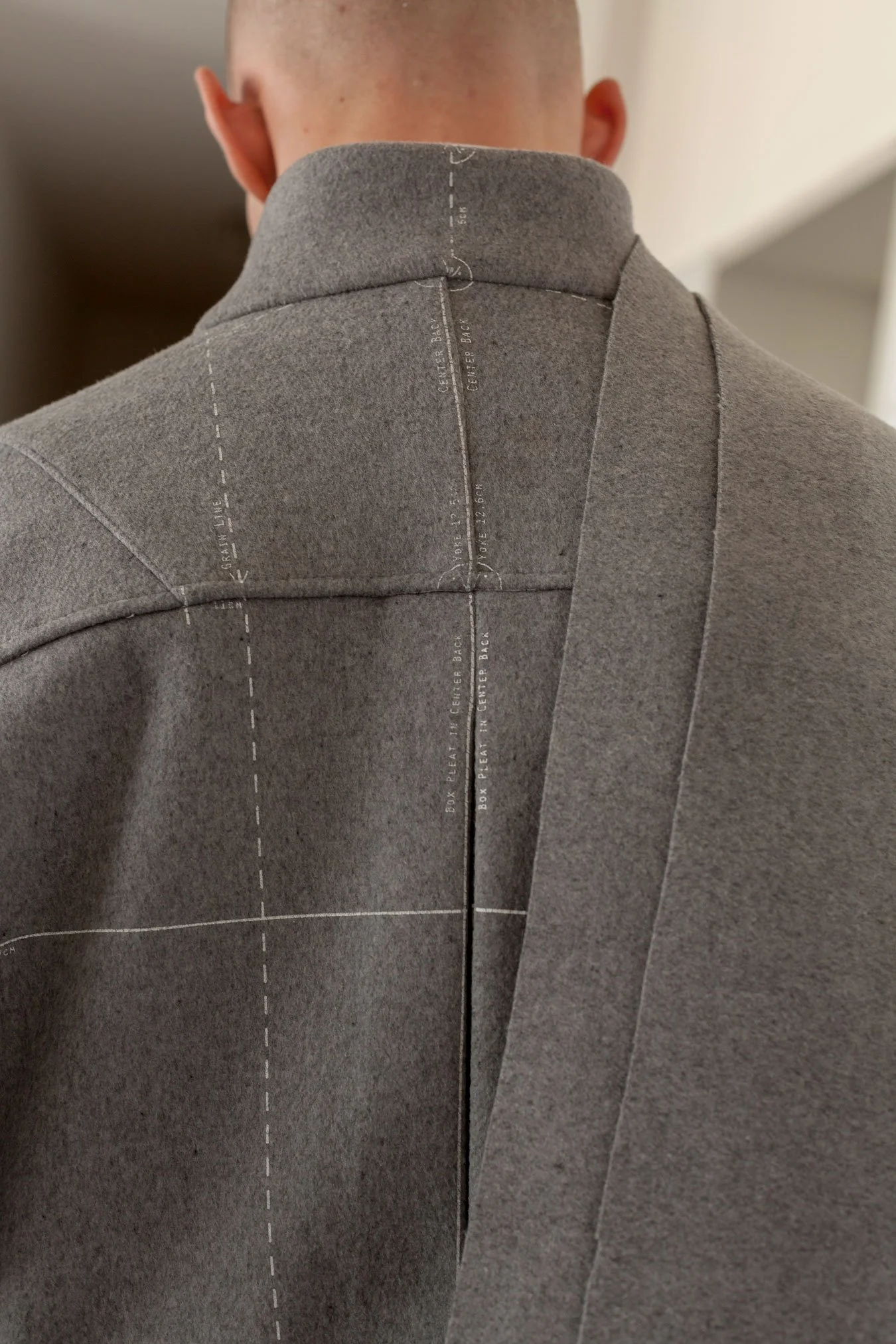 Nahaufnahme eines Mannes mit grauem Mantel, auf dessen Rücken Maßangaben und Anleitungen für die Schnittmustererstellung gedruckt sind.