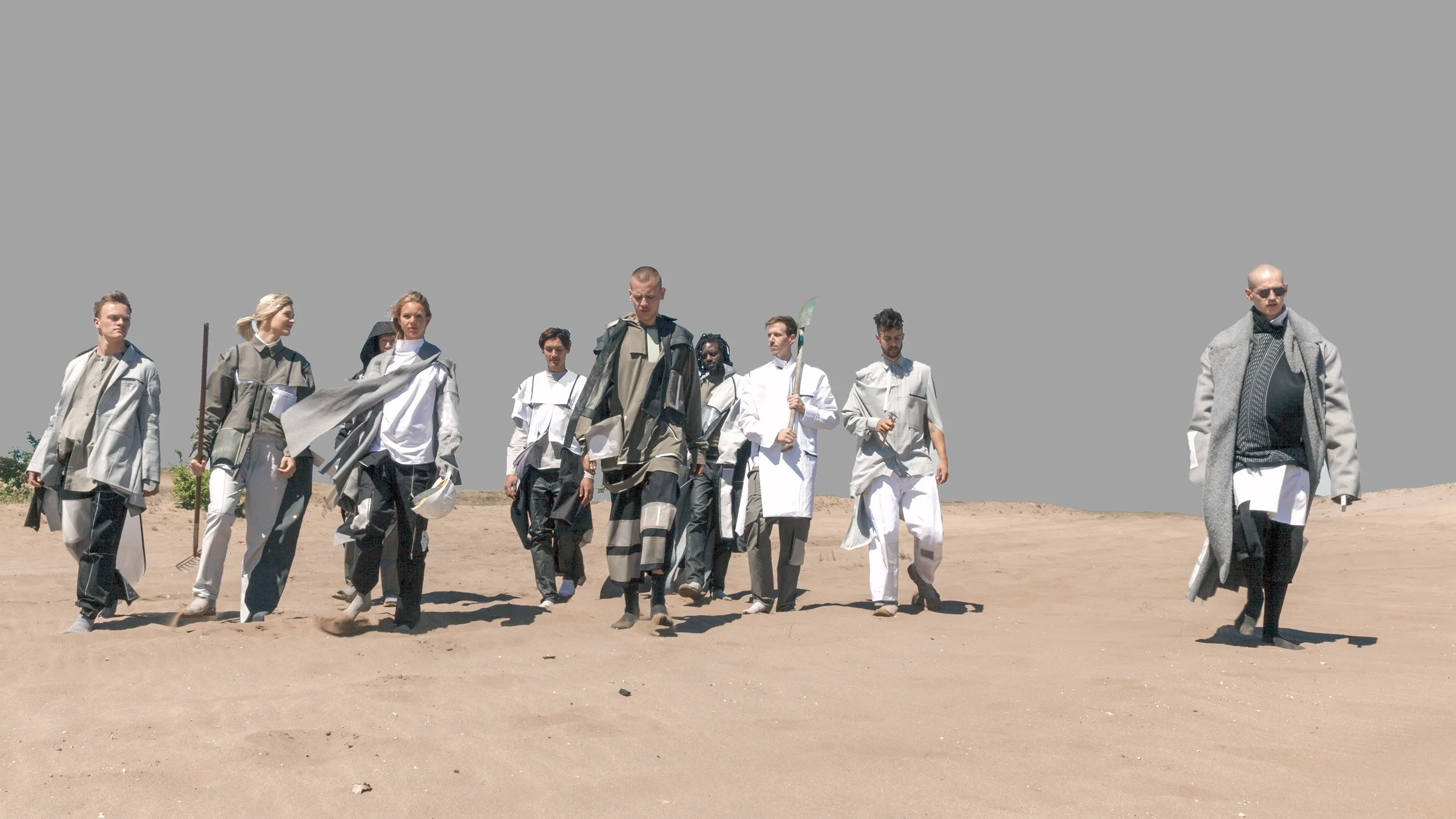Eine Gruppe von Menschen, die in weißen, grauen und schwarzen futuristischen und minimalistischen Outfits durch eine weite, kahle Wüstenlandschaft laufen.