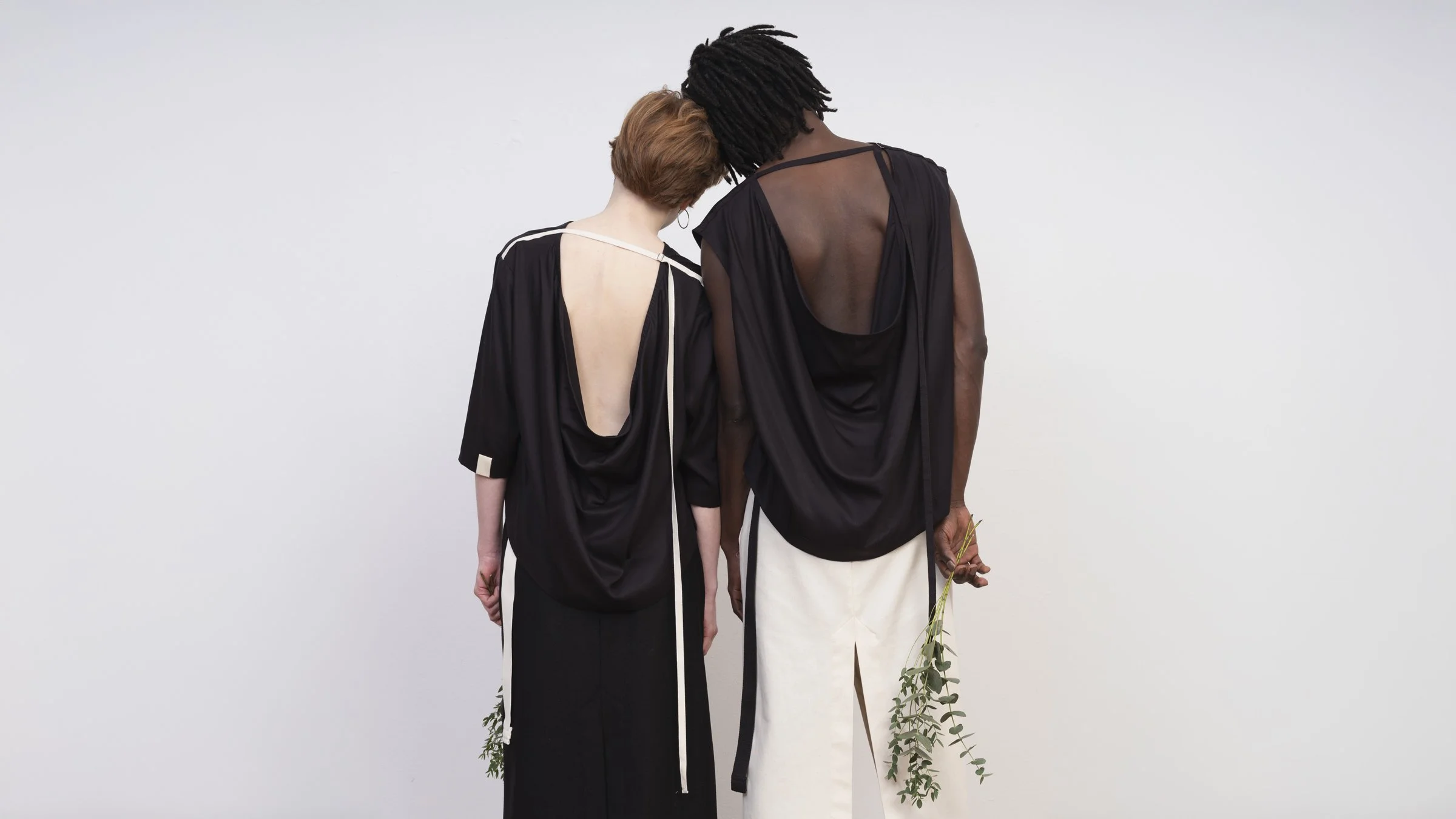 Zwei Personen stehen nebeneinander mit Rücken zur Kamera, beide tragen schwarze Wasserfall-Oberteile und Wickelröcke in Natur und Schwarz von studio.HIER. Sie halten kleine salbeizweige, vor einem weißen Hintergrund.
