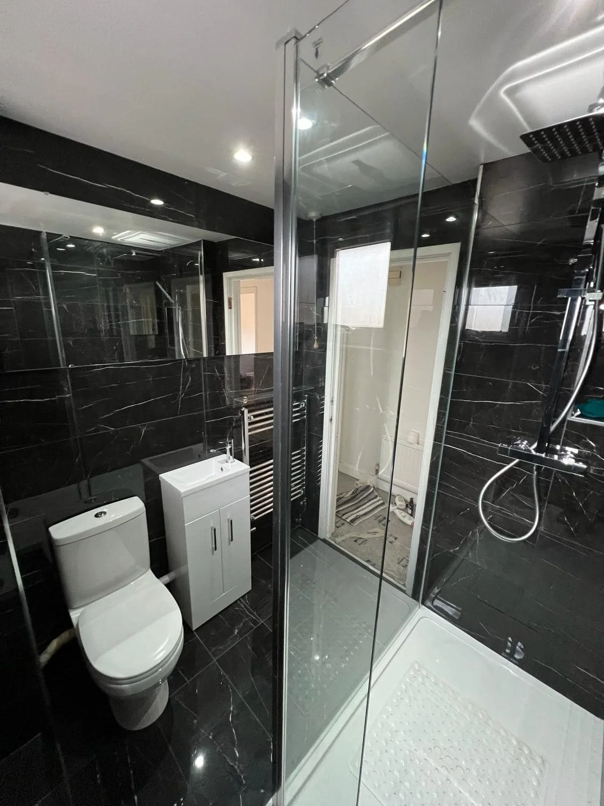 A modern bathroom with black marble walls and floor, a white toilet, a small white sink cabinet, a heated towel rail, and a shower with glass doors and a rainfall shower head.