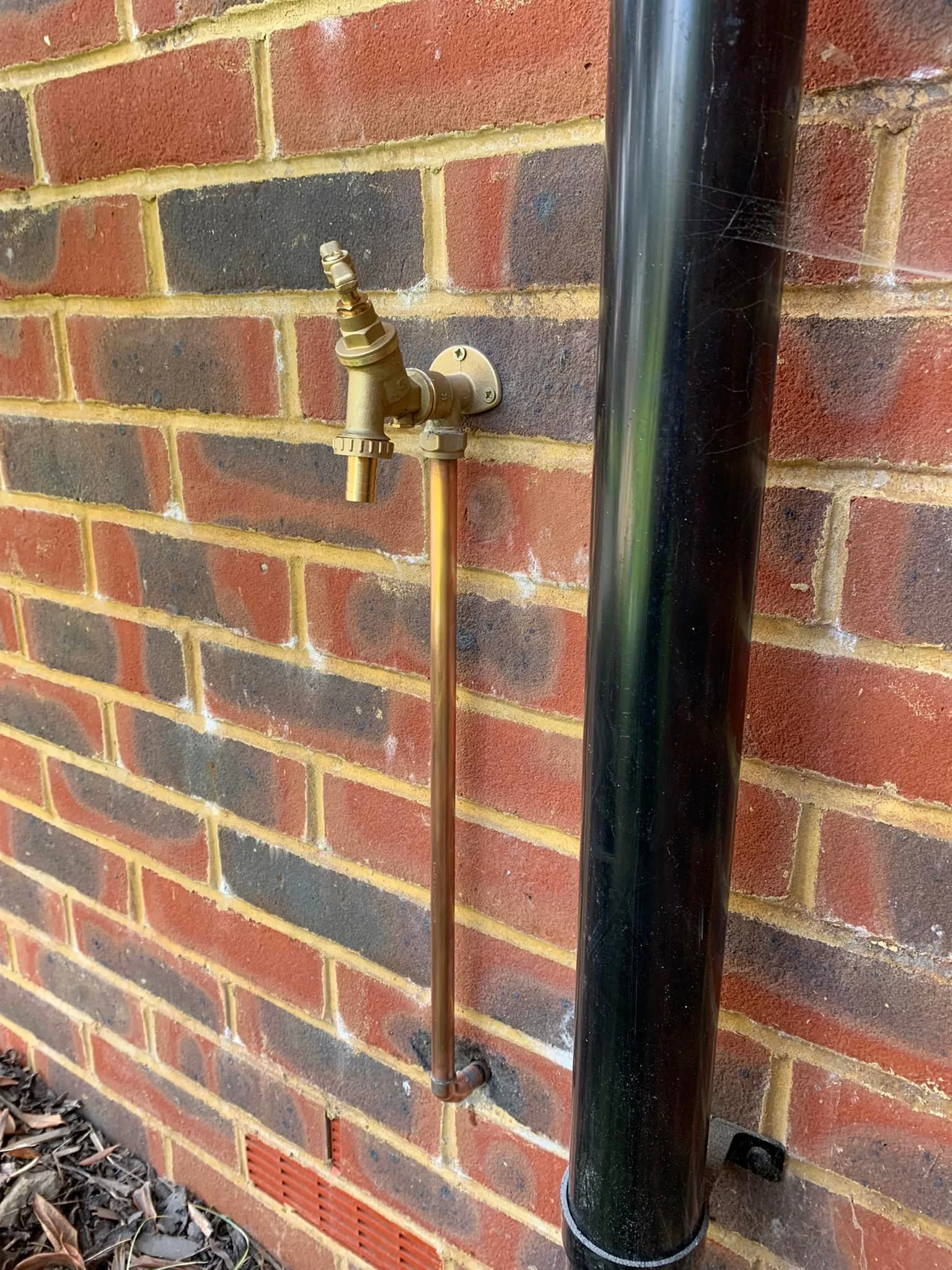An outdoor brass water faucet attached to a brick wall with a black drainpipe nearby.