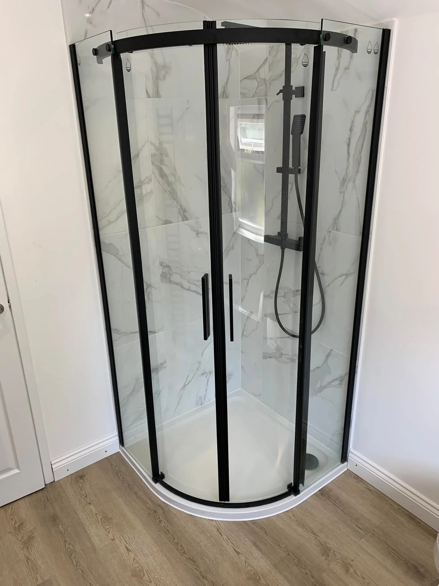 Corner shower with rounded glass doors and black framing, marble-patterned wall tiles, and a handheld showerhead inside.