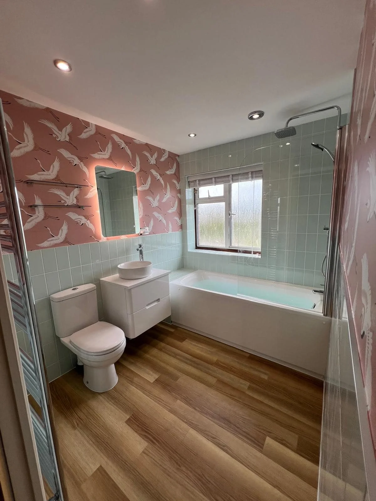 Bathroom with pink bird-patterned wallpaper, green tiled walls, a large window, a bathtub, a white sink on a floating cabinet, and a toilet, with wooden flooring and recessed ceiling lights.
