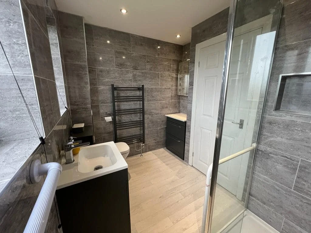 Modern bathroom with gray tiled walls, a white sink with a dark cabinet, a toilet, a black towel radiator, a small black cabinet with a white countertop, a glass shower enclosure, and a white door.
