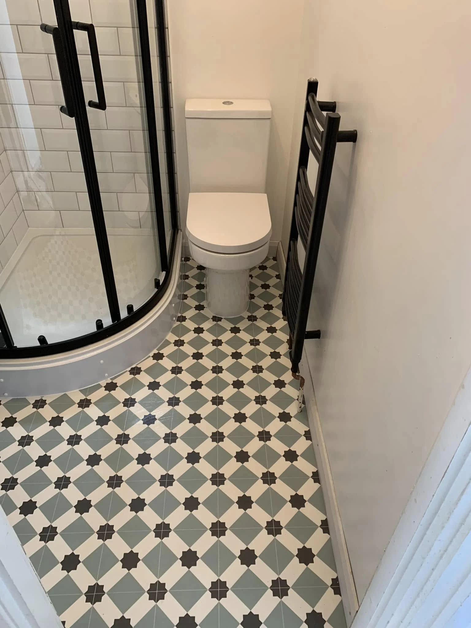 Small bathroom with patterned tile floor, toilet, corner shower with glass door, and a black towel rack.