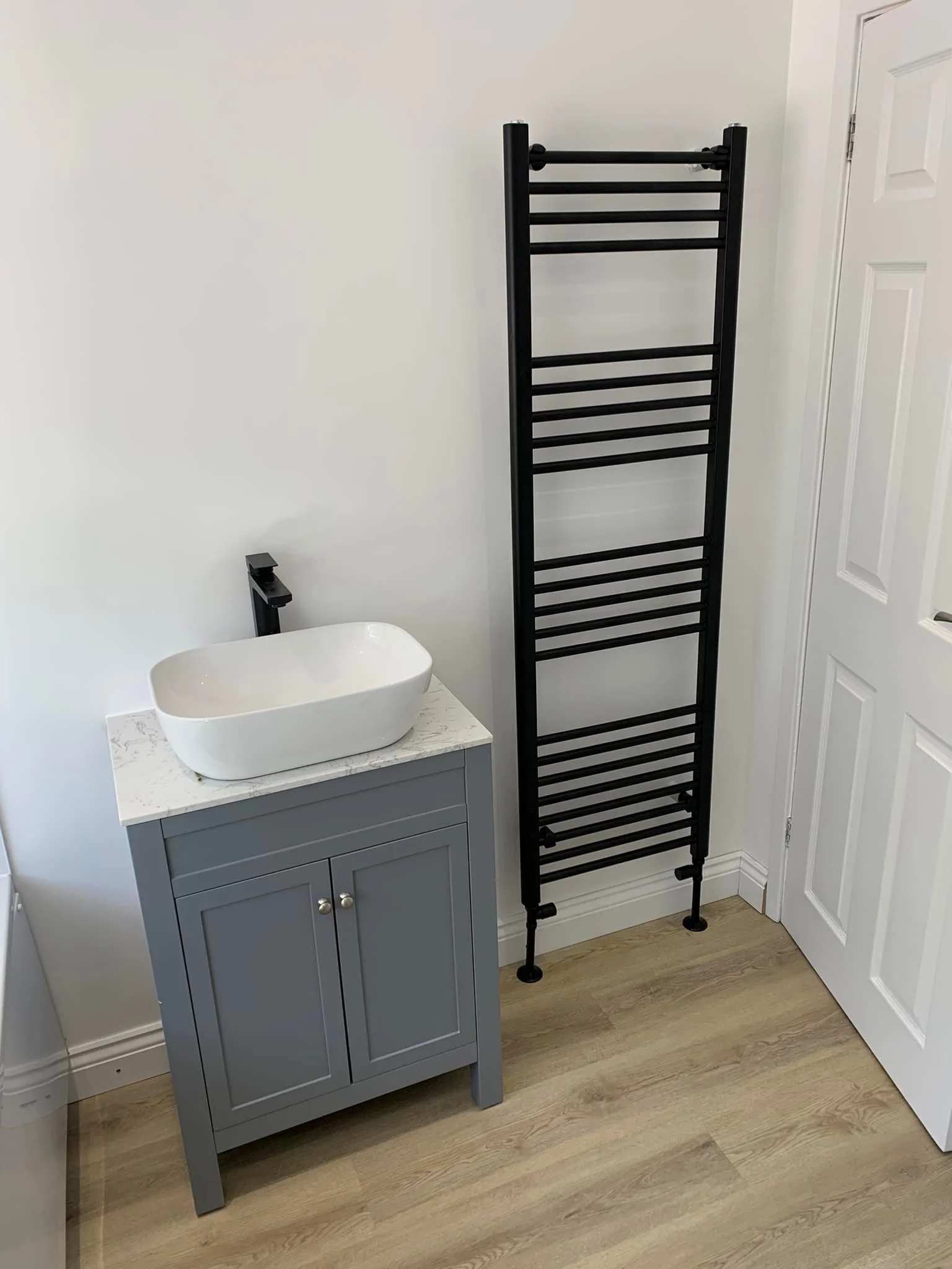 A bathroom corner with a gray cabinet holding a white vessel sink, a black faucet, and a black towel radiator against a white wall and wood flooring.