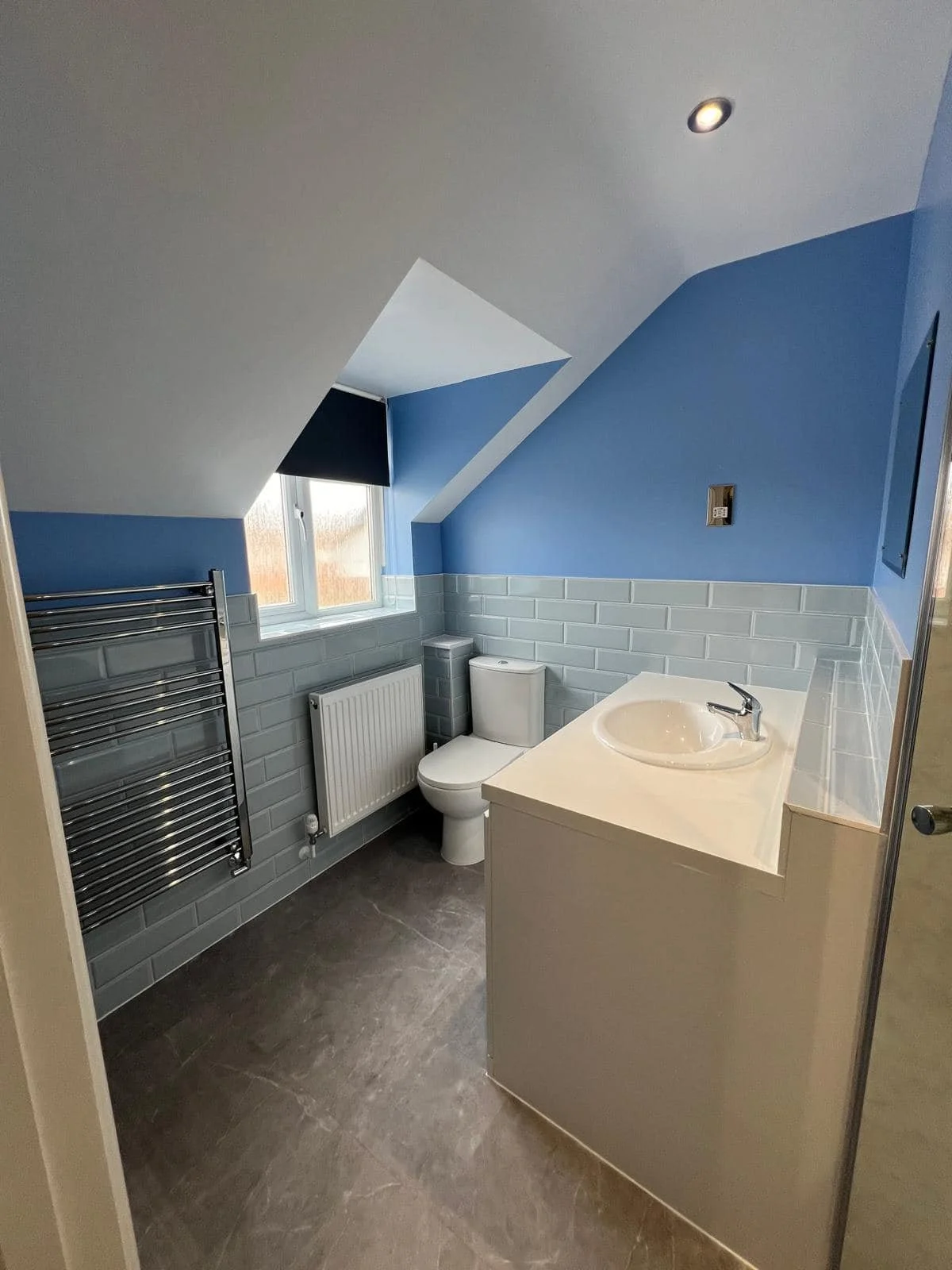 Small bathroom with blue walls and sloped ceiling, white toilet, white vanity with sink, window with frosted glass, and chrome towel radiator.