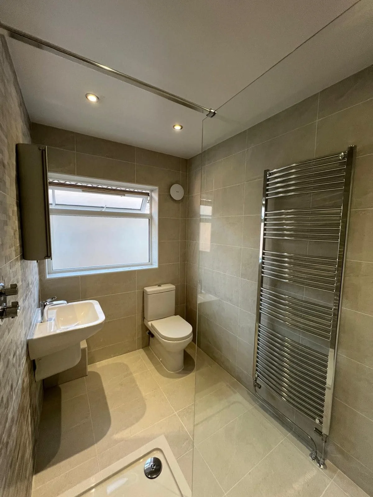 Small modern bathroom with beige tiled walls and floor, a window, a white toilet, a small white sink, a silver towel radiator, and recessed ceiling lights.