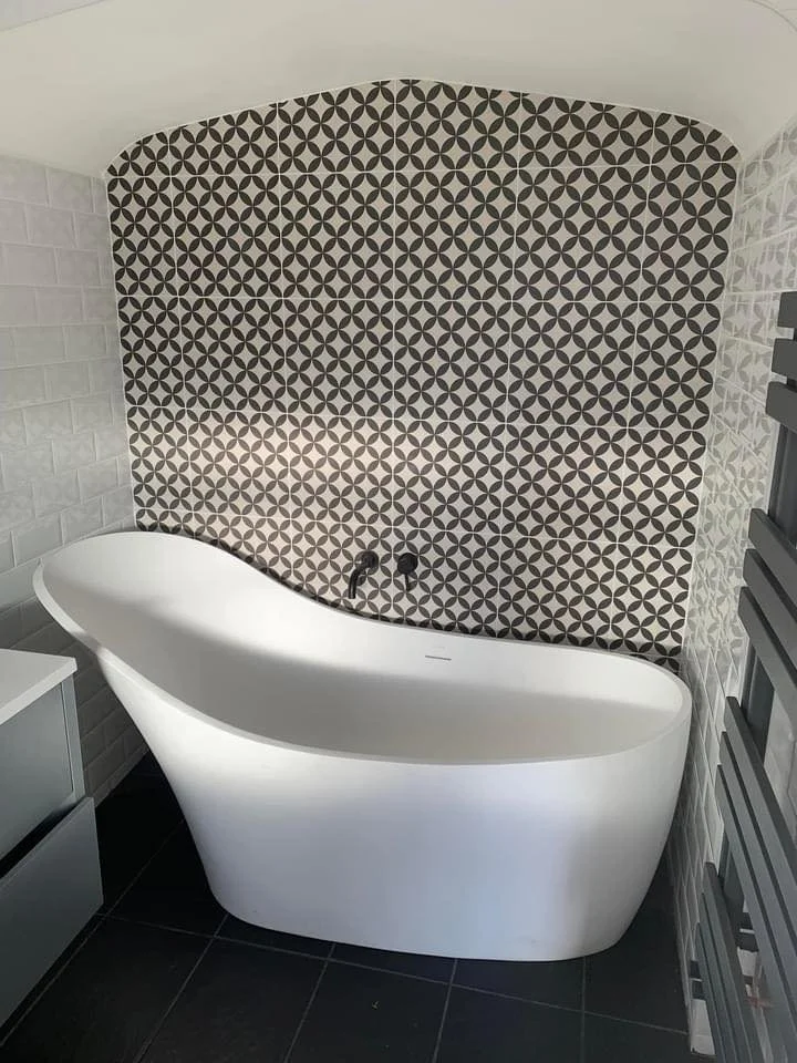 Modern bathroom with a white freestanding bathtub in front of a geometric patterned accent wall. To the right, there is a heated towel rail, and on the left, a small white cabinet. The floor is black tile.