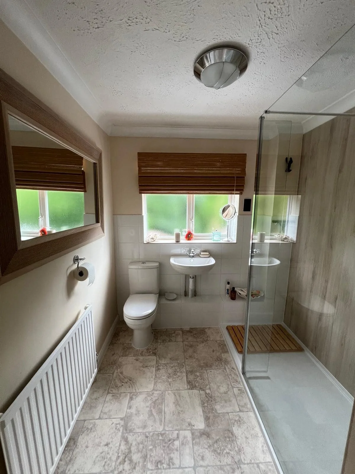 Bathroom with a toilet, small sink, shower area with wooden slatted floor, two small windows with brown blinds, mirror, and a radiator on the wall.