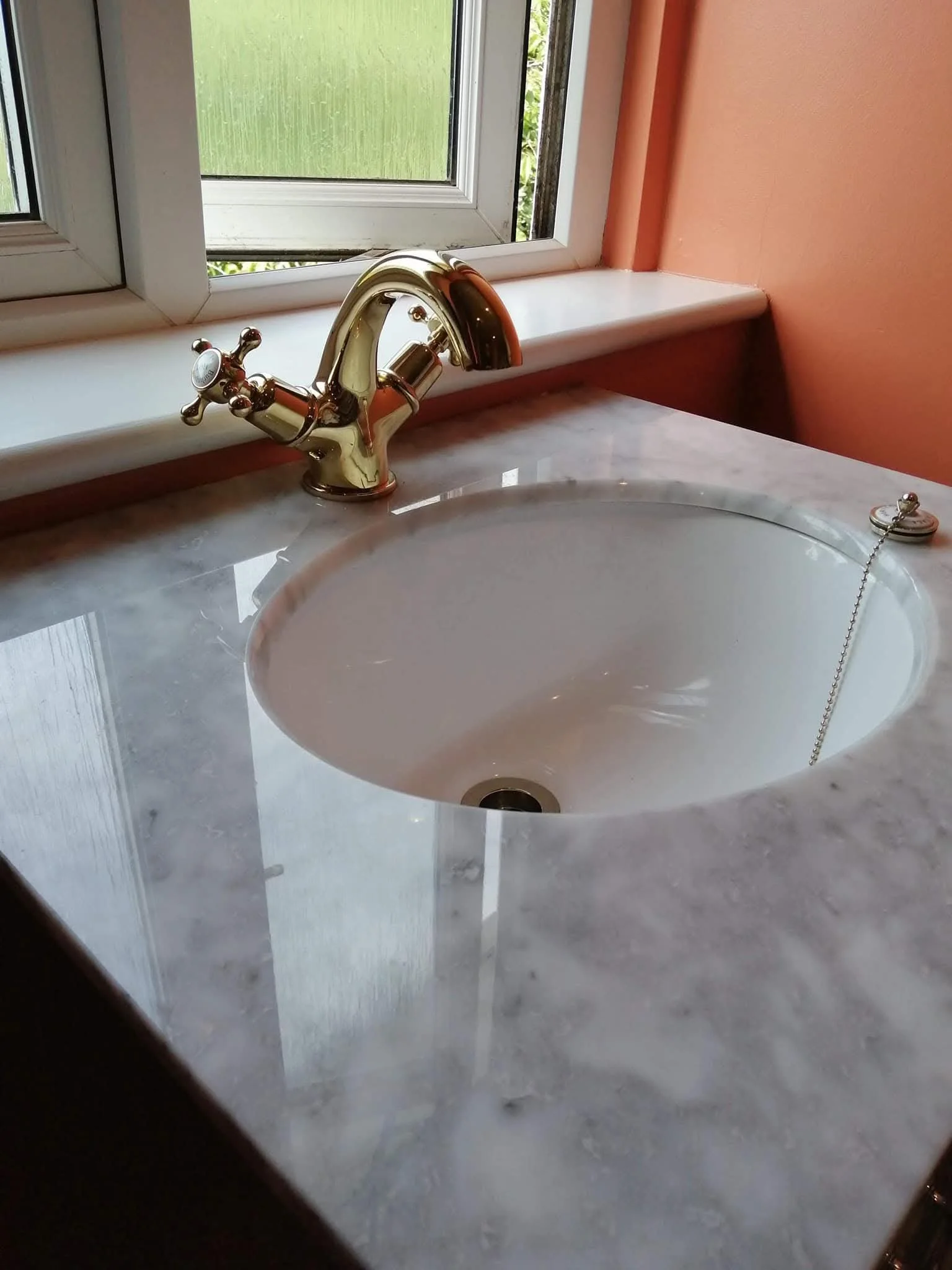 Close-up of a bathroom sink with a marble countertop, brass faucet, and a window above, with dark red and white walls.