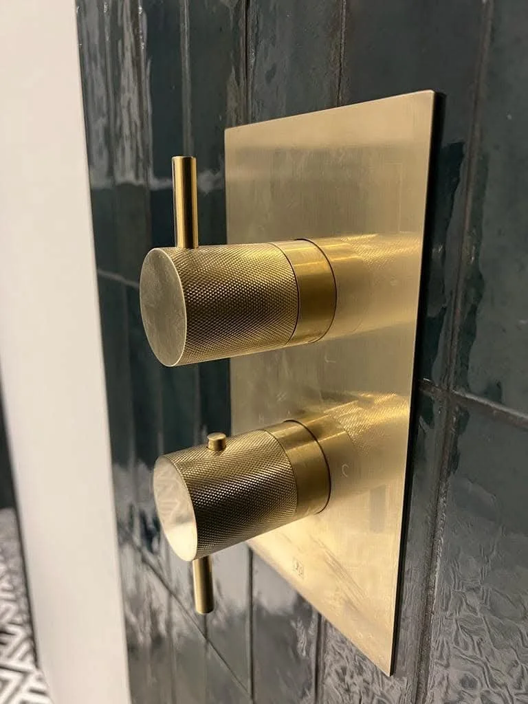 Close-up of a gold-colored bathroom faucet and control knobs mounted on a shiny metallic plate on a tiled wall.