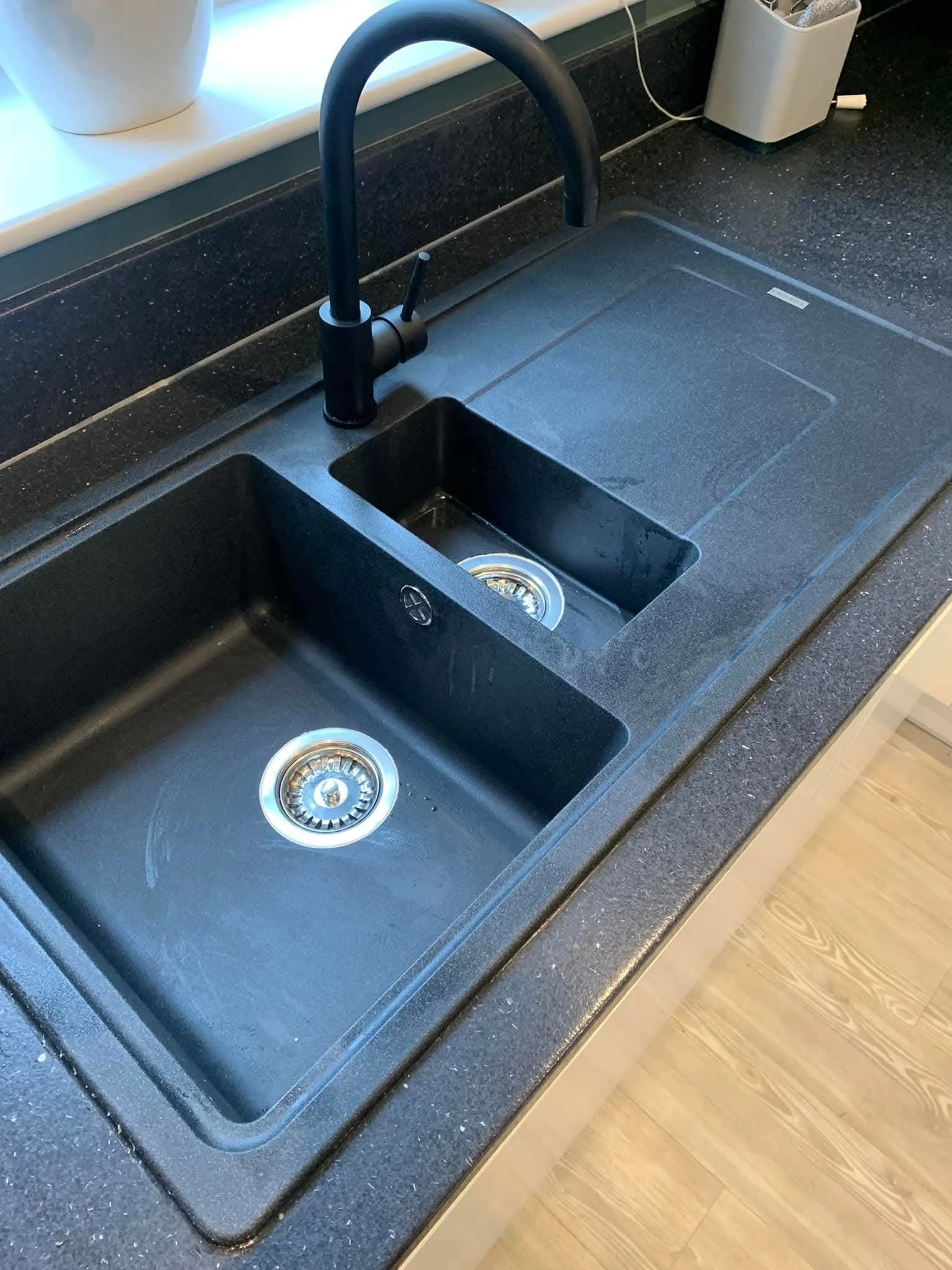 Black kitchen sink with two basins, one large and one small, and a black modern faucet on a dark granite countertop in a kitchen. There is a potted plant on the windowsill and a white container with some utensils nearby.