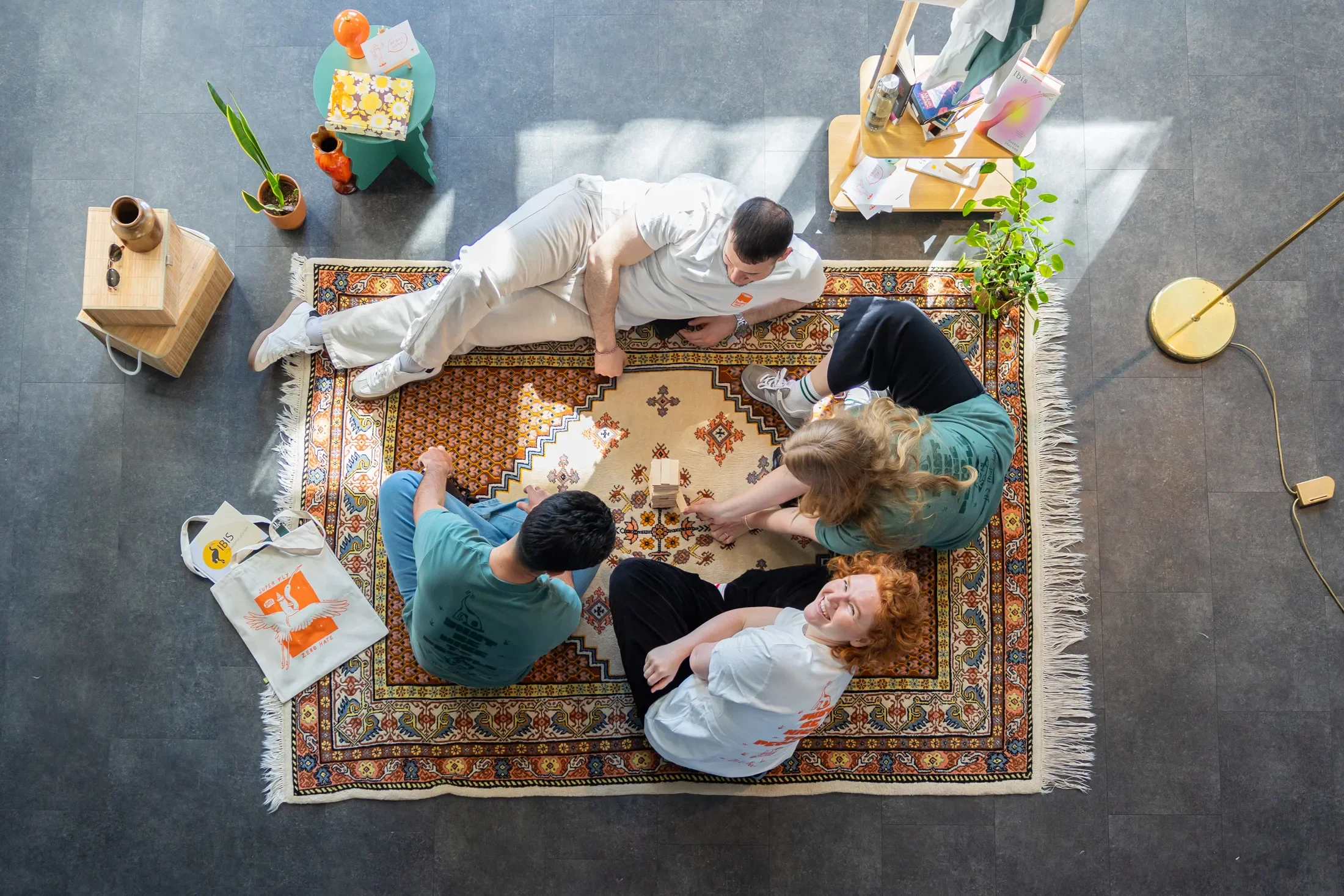 Eine Gruppe von Menschen, die auf einem bunten Teppich sitzen, unter einem Dach, mit Pflanzen, Büchern und einem Kabel. Es gibt Sonnenlicht und eine entspannte Atmosphäre.
