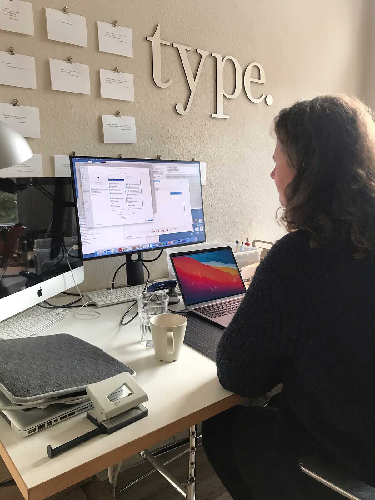 Superidee Mitarbeiterin Nina im Home Office an ihrem Computer und Laptop sitzend. Sie ist nur von seitlich angeschnitten zu sehen. Hinter ihrem Monitor ist eine Pinnwand mit den Buchstaben "type" zu sehen.