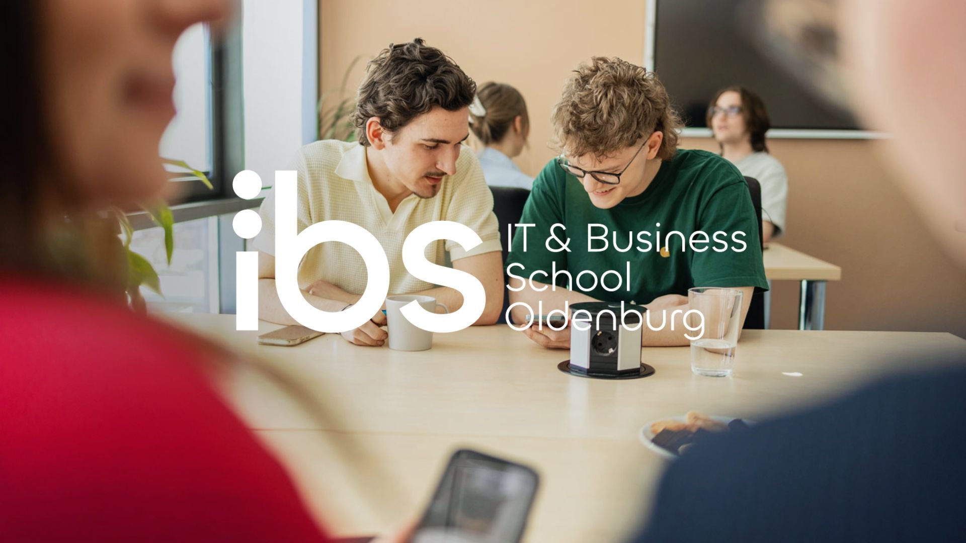 Zwei junge Männer schauen auf ein Smartphone. Im Vordergrund sind die Schultern einer Person in Rot und einer in Schwarz. Im Hintergrund sind weitere Personen in einer Schulklasse. Es ist ein Lehr- und Lernumfeld, mit einem Whiteboard und einem Fenster im Raum.