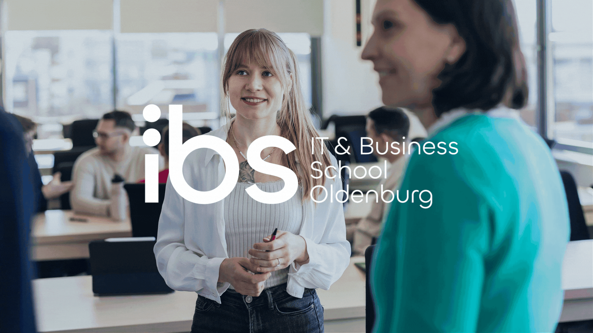 Frauen sprechen in einem Klassenzimmer, in dem mehrere Studierende an Tischen sitzen, mit großem Fenster im Hintergrund, Textüberlagerung mit Logo und Information zum IBS Oldenburg.