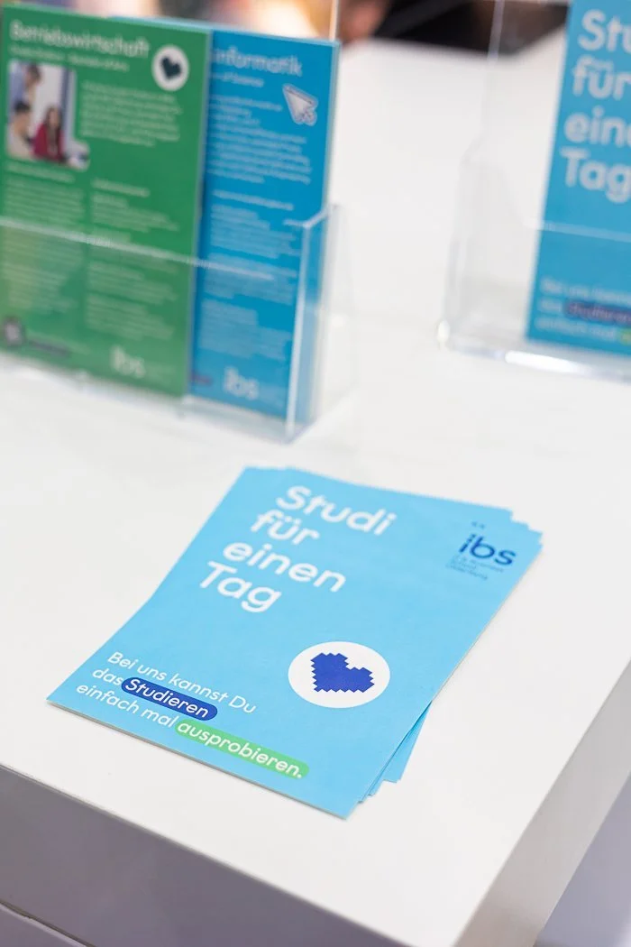 Flyer mit blauer Überschrift 'Studi für einen Tag', die zum Ausprobieren einlädt, auf einem weißen Tisch, im Hintergrund Informationsbroschüren in grüner und blauer Verpackung.