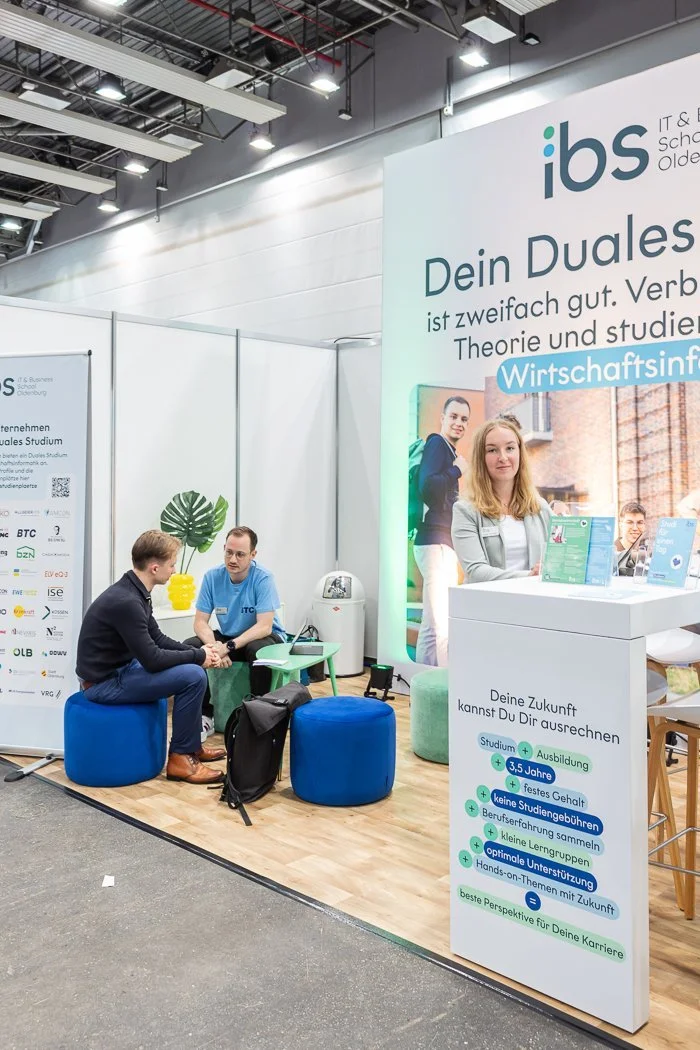 Messestand der IBS (Internationale Business School) mit einem großen Poster, das die Vorteile des Dualen Studiums hervorhebt, und zwei jungen Männern im Gespräch sowie einer jungen Frau am Empfangstresen.
