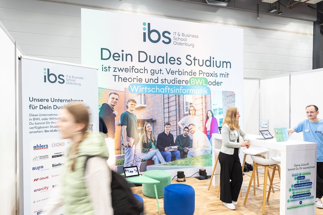 Messestand des IT & Business School Oldenburg mit großen Plakaten, auf denen das duale Studium beworben wird, Menschen sprechen mit einem Vertreter, Sitzgelegenheiten und Infomaterial.