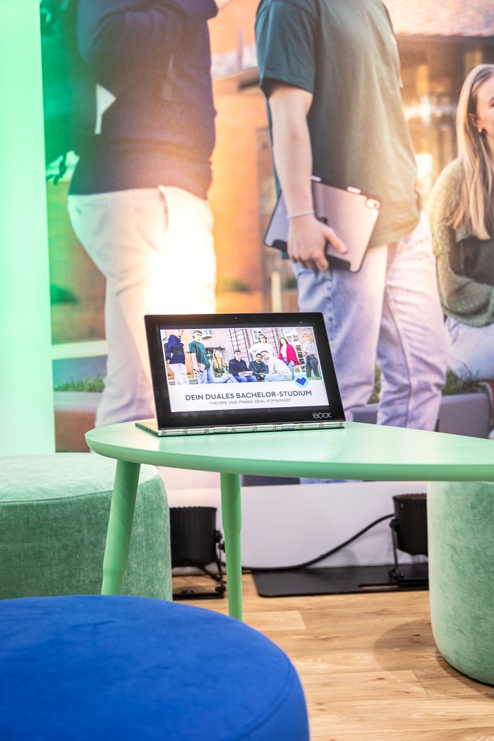 Ein Tablet auf einem grünen Tisch zeigt eine Gruppe von Menschen, die an einem Event teilnehmen. Im Hintergrund ist eine große Wand mit einem Foto von mehreren Menschen, die im Freien sitzen oder stehen. Text auf dem Tablet liest: "DEIN DUALES BACHELOR-STUDIUM".