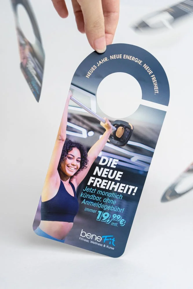Werbeanhänger für Bene Fit Fitnessstudio mit einem lächelnden jungen Frau im Sportoutfit, die mit einer Hantel trainiert. Text auf dem Anhänger: 'Die neue Freiheit! Jetzt monatl. kündbar, ohne Anmeldegebühr! 19,99 € monatlich.'