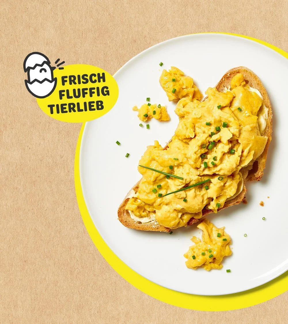 Ein Teller mit Rührei auf Vollkornbrot, garniert mit Schnittlauch, auf einem weißen Teller mit gelber Umrandung, neben einem Logo mit einem Ei und dem Text 'Frisch Fluffig Tierlieb'