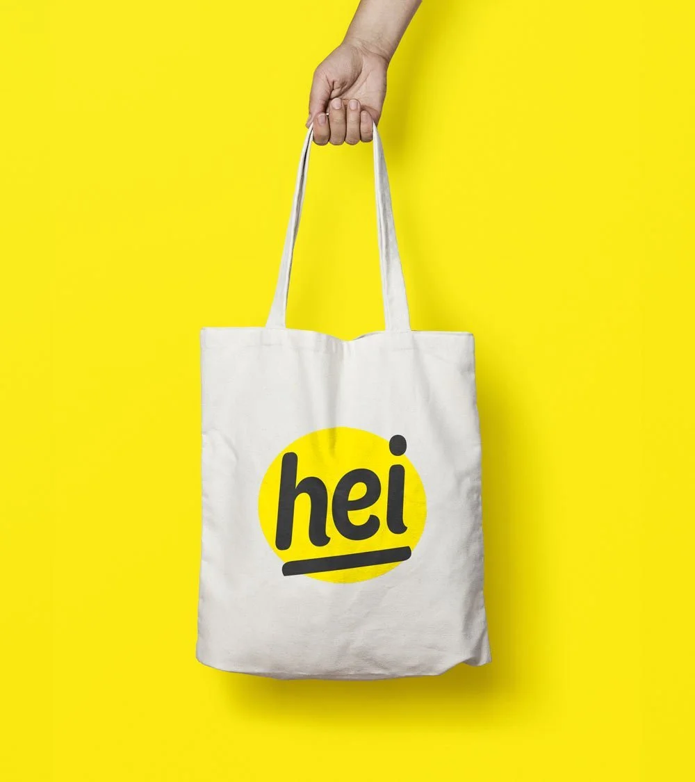 Hand hält einen weißen Stoffbeutel mit dem schwarzen Schriftzug "hei" vor einem gelben Hintergrund.