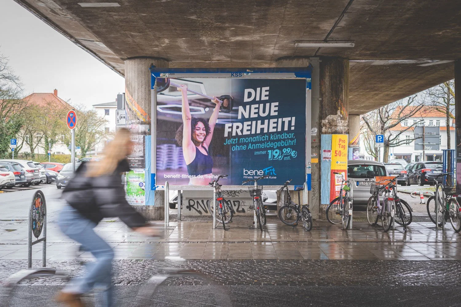 Blick auf eine Unterführung mit Fahrradständern und mehreren Fahrrädern, ein einfahrender Fahrradfahrer im Vordergrund, eine große Werbetafel an der Wand der Unterführung. Die Werbung zeigt eine Frau, die eine Hantel hebt, mit dem Text 'DIE NEUE FREIHEIT! Jetzt monatlich kündbar, ohne Anmeldegebühr! 19,99 € monatlich'. Im Hintergrund sind parkende Autos und baufällige Häuser sichtbar.
