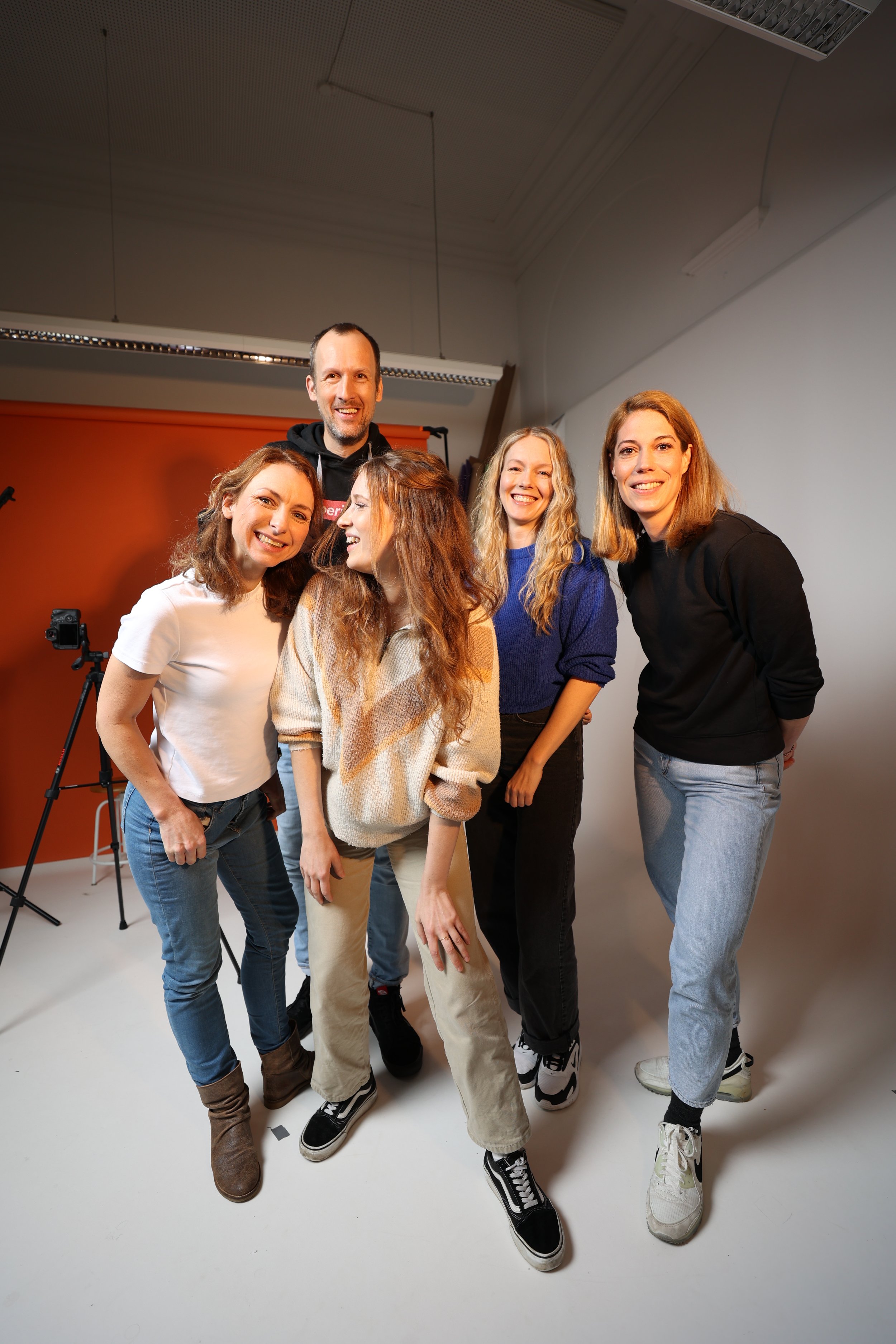 Fünf junge Menschen posieren lachend in einem Fotostudio, einige tragen sportliche Kleidung und Sneakers, im Hintergrund ist ein rot-orangefarbener Hintergrund und Fototechnik sichtbar.