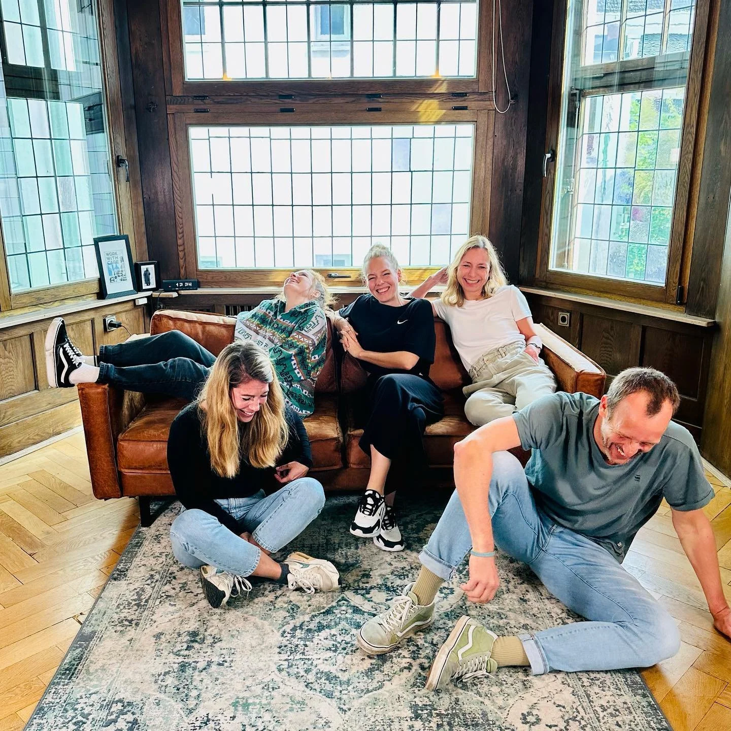 Eine Gruppe von fünf Menschen sitzt und liegt lachend auf einem Sofa und Boden in einem Holzhaus mit großen Fenstern im Hintergrund.