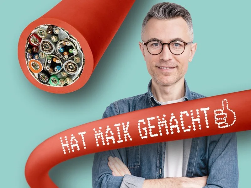 Ein Mann mit Brille steht vor einer türkisfarbenen Wand, umgeben von einem roten Kabel. Das Kabel hat einen Schriftzug in Form von Leuchtdioden, der lautet 'HAT NACK GEMACHT'.