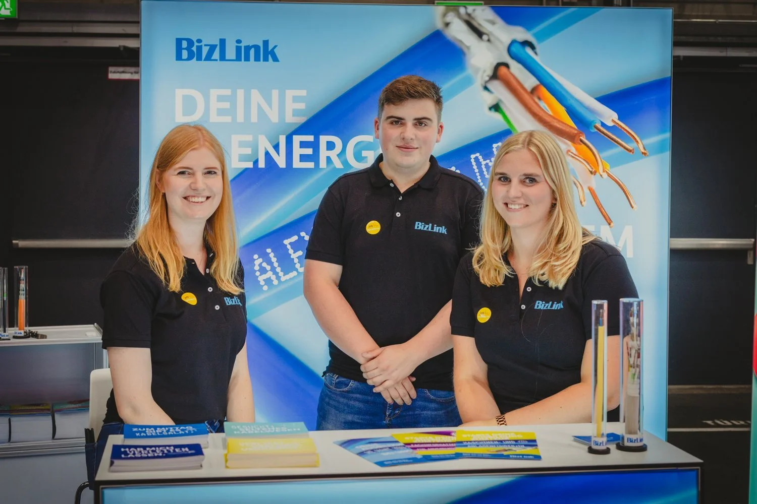 Drei Personen stehen hinter einem Stand mit Stromkabeln und Flyern, im Hintergrund ein Banner mit der Aufschrift 'BizLink' und 'Deine Energie'.