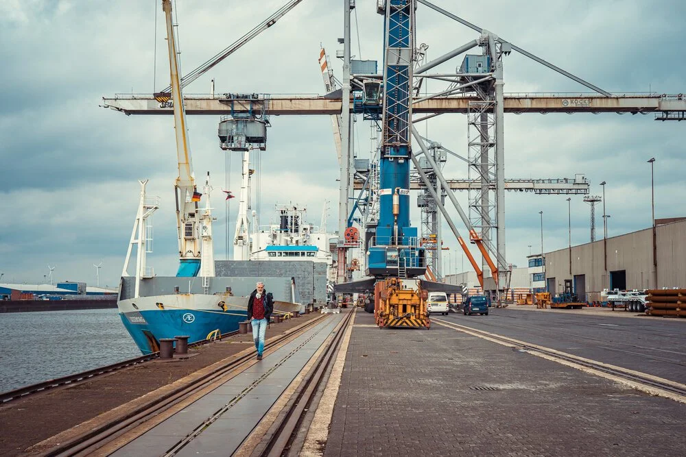 Blick auf einen Hafen mit großen Kränen und einem großen Frachtschiff, das am Ufer liegt. Es gibt auch eine Person, die neben den Bahngleisen läuft.