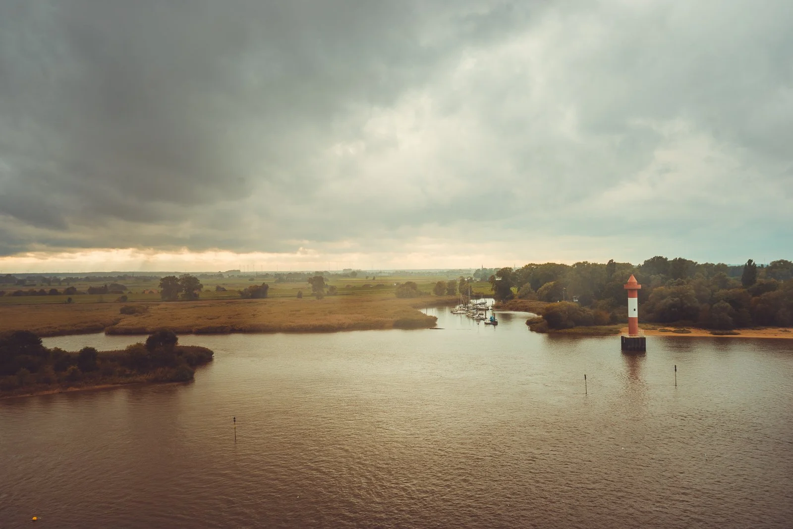 Eine ruhige Wasserstraße mit einem Leuchtturm und Segelbooten, umgeben von grünen Hügeln und Bäumen bei bewölktem Himmel.