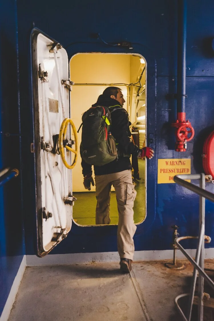 Eine Person mit Rucksack steigt durch eine lukende Tür in ein Schiff. Die Tür ist mit einem Dickicht aus Schläuchen und Ventilen umgeben, und es gibt eine Warnschilder mit der Aufschrift "Warning Pressureized".