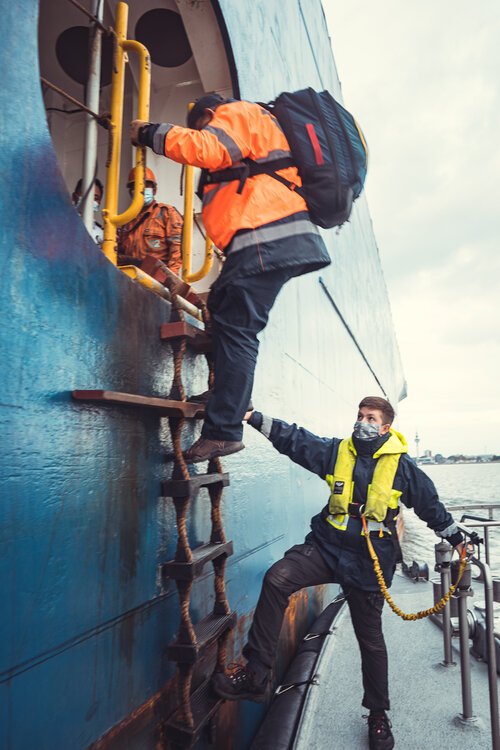Zwei Menschen beim Einsteigen in ein Schiff, eine hilft der anderen, während sie eine Leiter benutzen, alle tragen Wetterkleidung und eine Person trägt eine Sicherheitsweste.