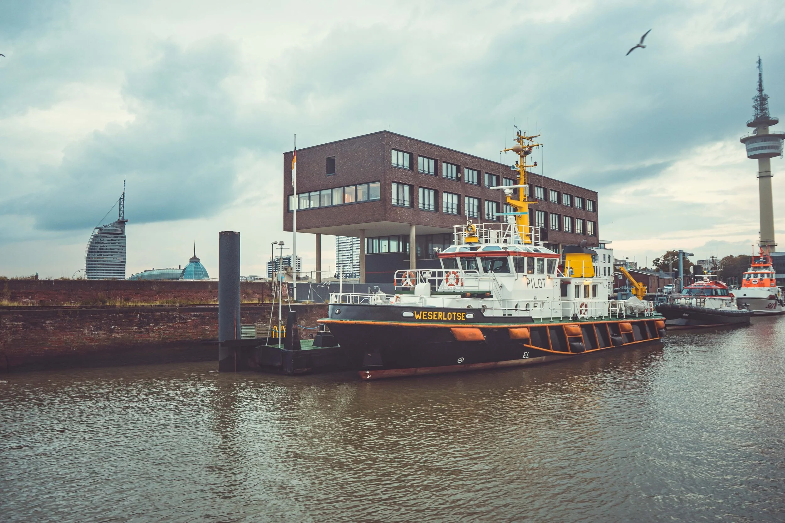 Boote im Hafen vor modernen Gebäuden und einem Turm, bei bewölktem Himmel.