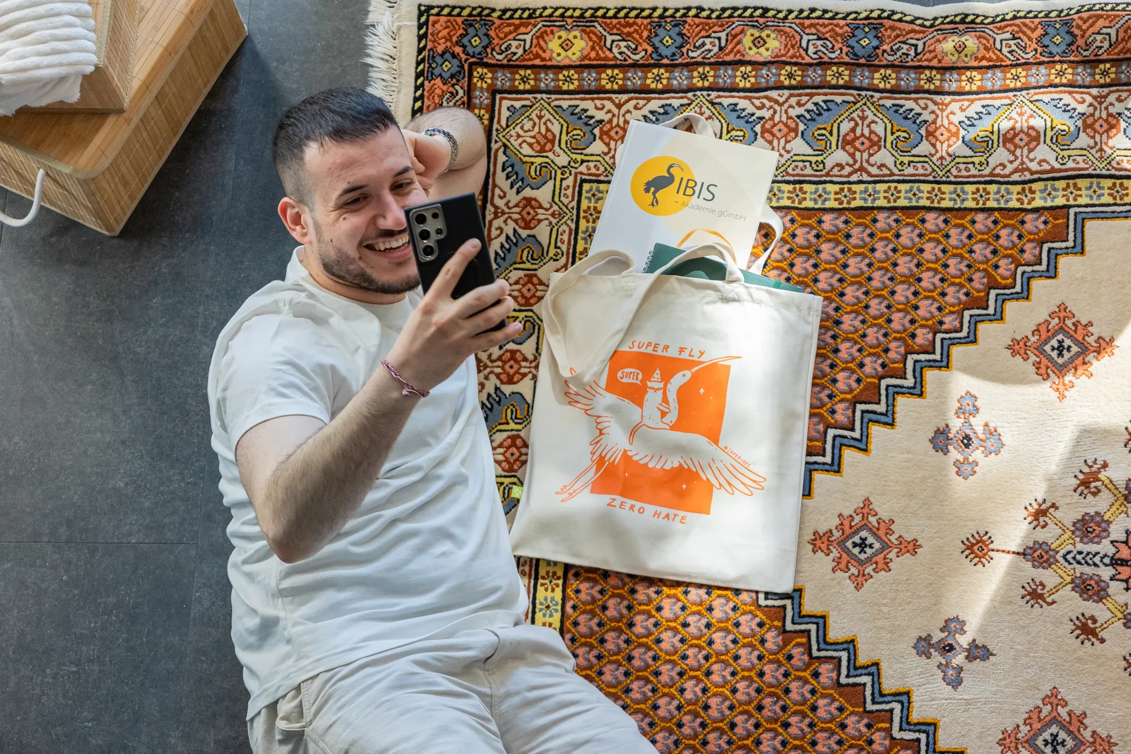 Person liegt entspannt auf einem Teppich neben der Super-Tasche, mit IBIS e. V. Notizbuch und fotografiert mit ihrem Smartphone – Behind the Scenes aus dem Superidee Werbeagentur Online-Shop.