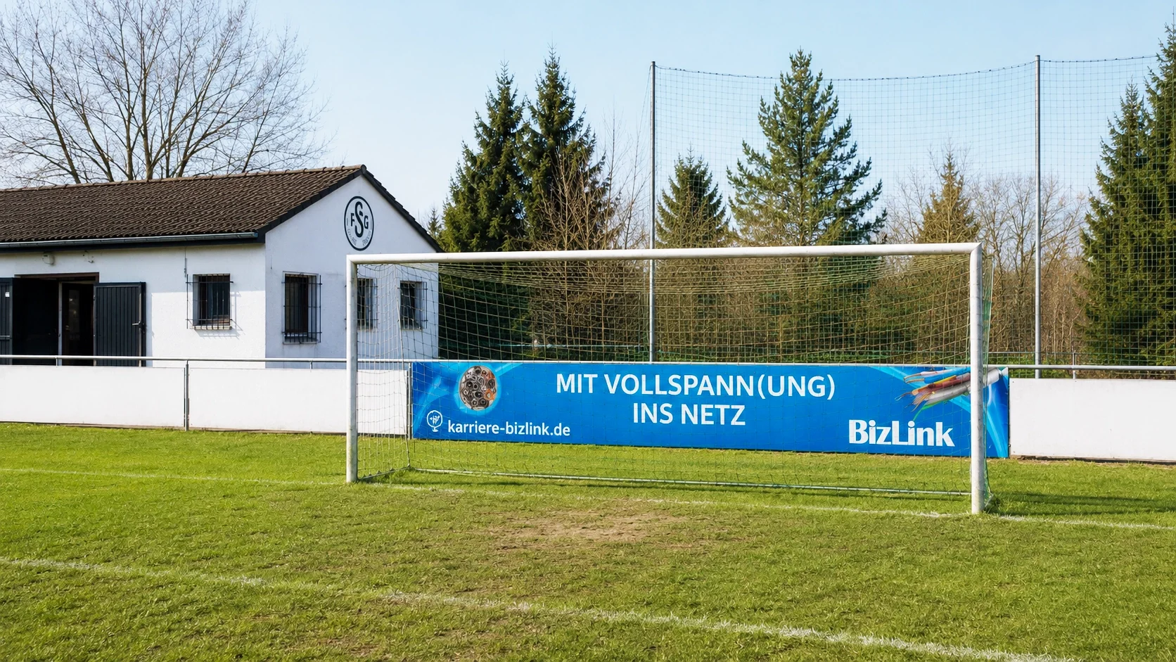Fußballfeld mit Tor, das eine Werbung mit dem Text 'Mit Vollspann(ung) ins Netz' und der Webseite karriere-bizlink.de trägt. Im Hintergrund ein weißes Gebäude mit dem Logo FSG
