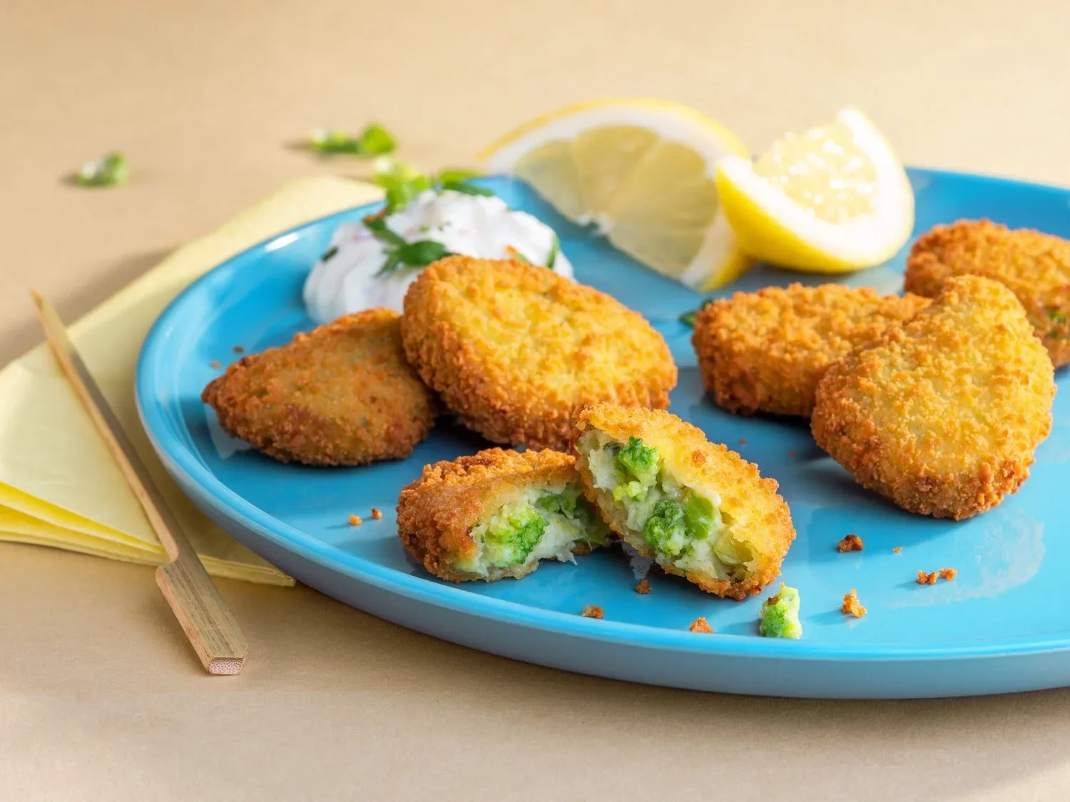 Frittierte Eierfrikadellen mit Erbsenfüllung auf einem blauen Teller, dazu Zitronenscheiben und Zitronenviertel, andeutende Zitronenspalten, Creme mit Kräutern