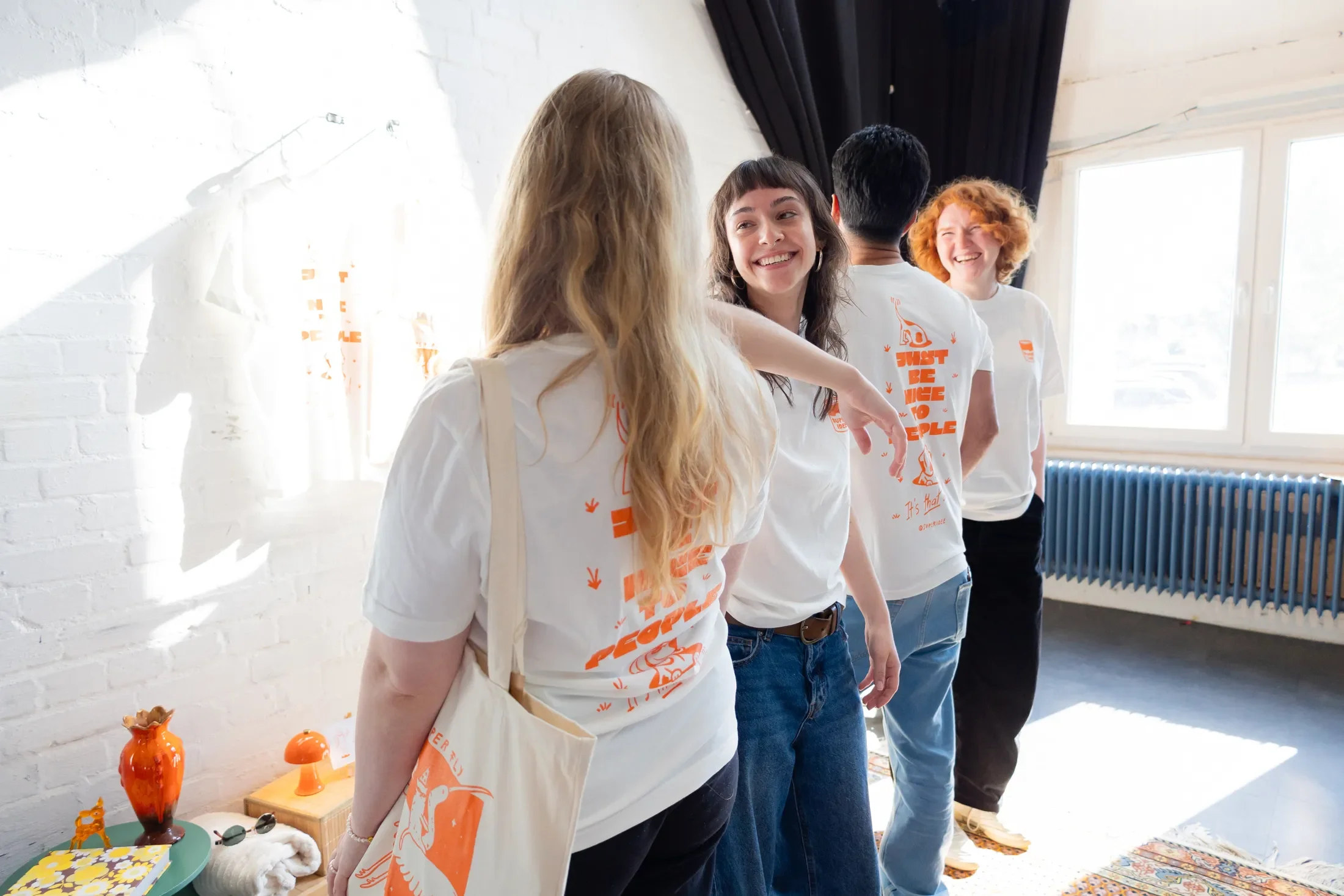 Menschen stehen in einer Gruppe in einem hell erleuchteten Raum, einige lachen und führen ein Gespräch. Sie tragen weiße T-Shirts mit orangefarbenem Druck. Im Vordergrund ist eine Frau mit langen, gewellten blonden Haaren, die einen Beutel trägt. Im Hintergrund sind Fenster, Vorhänge und Heizkörper sichtbar, sowie Dekorationsgegenstände auf einem Tisch.